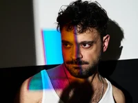 Ein Mann in einem weißen Tanktop steht im Schatten. Farbige Lichtprojektionen fallen auf sein Gesicht und den Hintergrund.