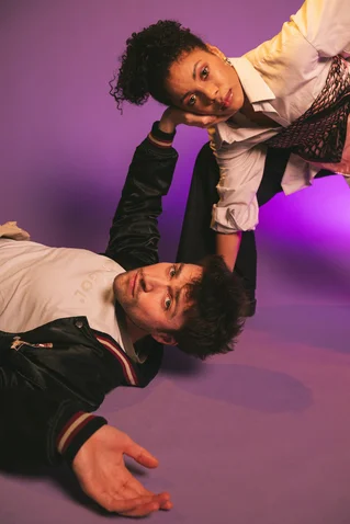 A man lies on his back on a purple floor, while a woman stands over him, posing elegantly.