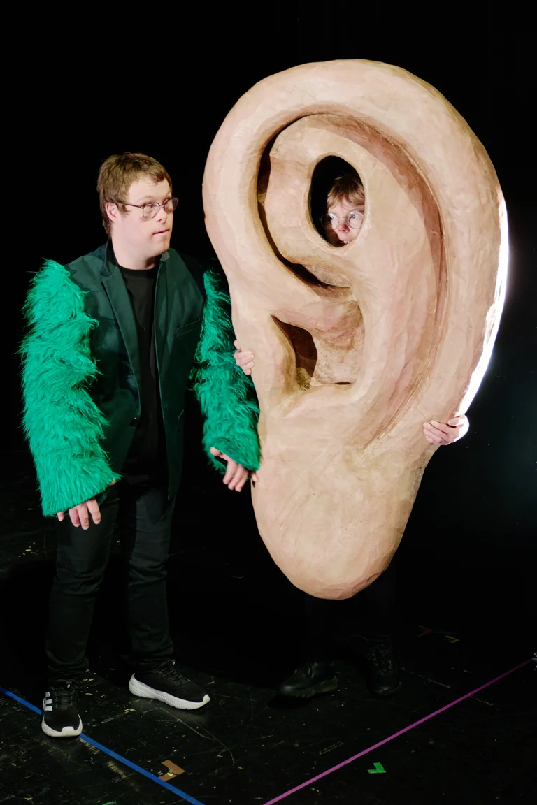 Ein Mann in grüner Jacke steht neben einer großen Ohren-Requisite. Eine Person hält das Ohr von hinten fest.