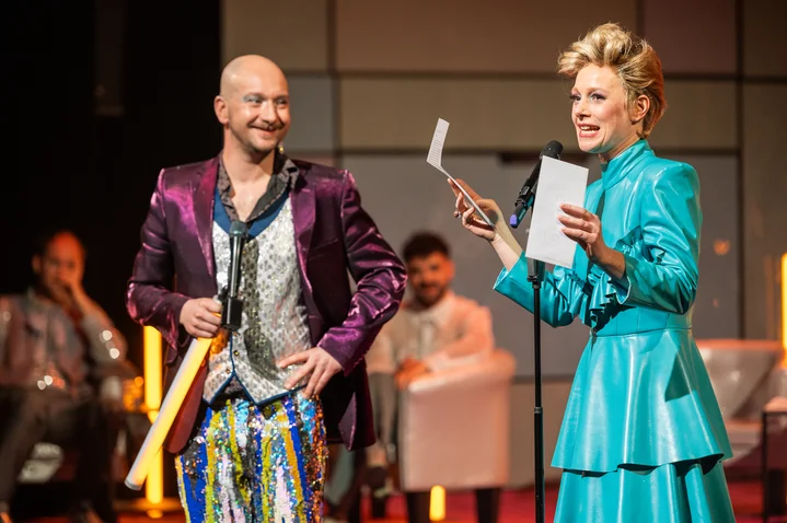 Two people on a stage in colorful outfits, holding papers. Audience members sit in the background, observing.