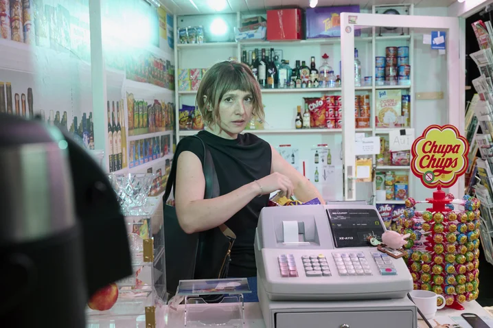 A woman stands behind the counter in a small shop, surrounded by snacks and drinks. A lollipop display is visible.