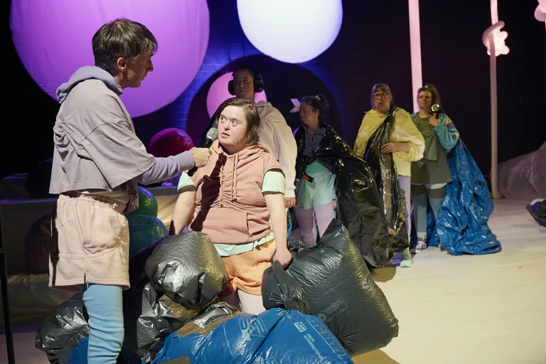 Eine Theateraufführung mit mehreren Personen auf der Bühne. Einige halten blaue Taschen, während sie miteinander interagieren.