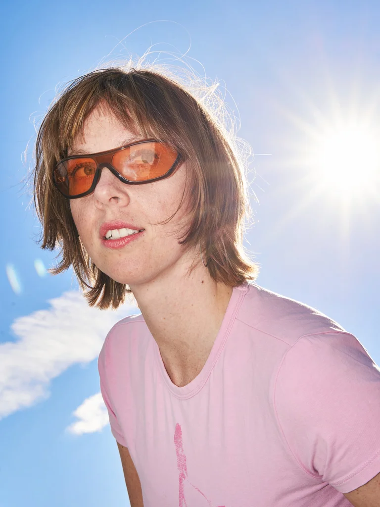 Person mit braunem Haar, Sonnenbrille und rosa T-Shirt vor blauem Himmel und Sonne im Hintergrund.