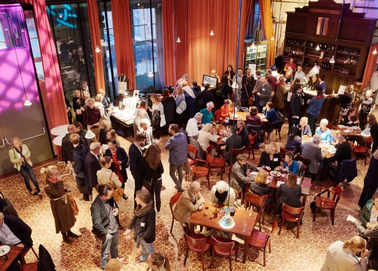 Eine gut besuchte Veranstaltung in einem eleganten Raum, mit Menschen, die sich an Tischen unterhalten und eine Bar im Hintergrund.