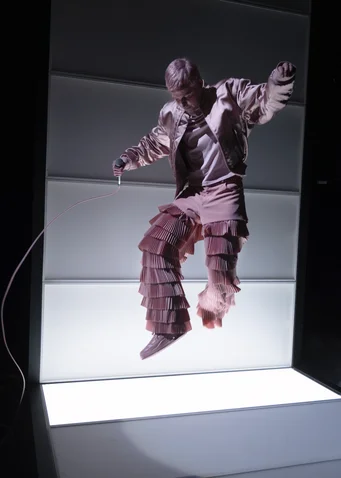 A person in a striking, layered outfit jumps dynamically on a lit stage. The background is dark.