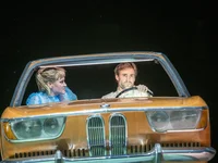 Two people in a car on stage, under dramatic lighting, with intense expressions, depicting a theatrical scene.