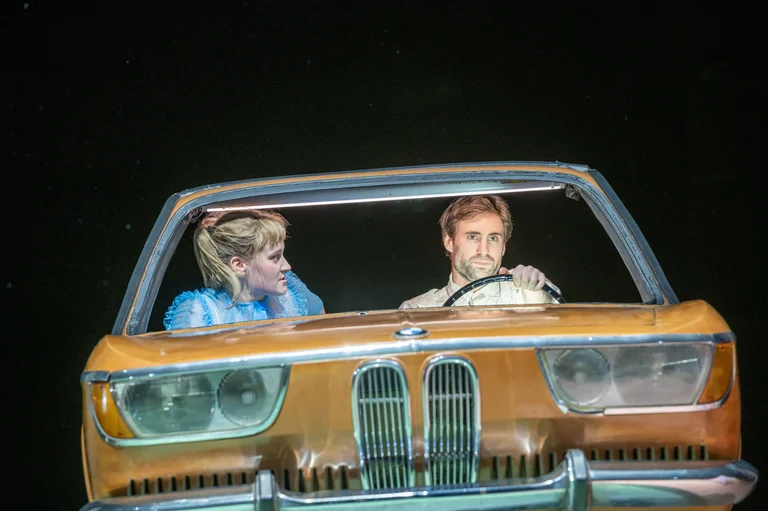 Zwei Personen in einem Auto auf der Bühne, mit dramatischer Beleuchtung und intensiven Ausdrücken, die eine theatralische Szene darstellen.