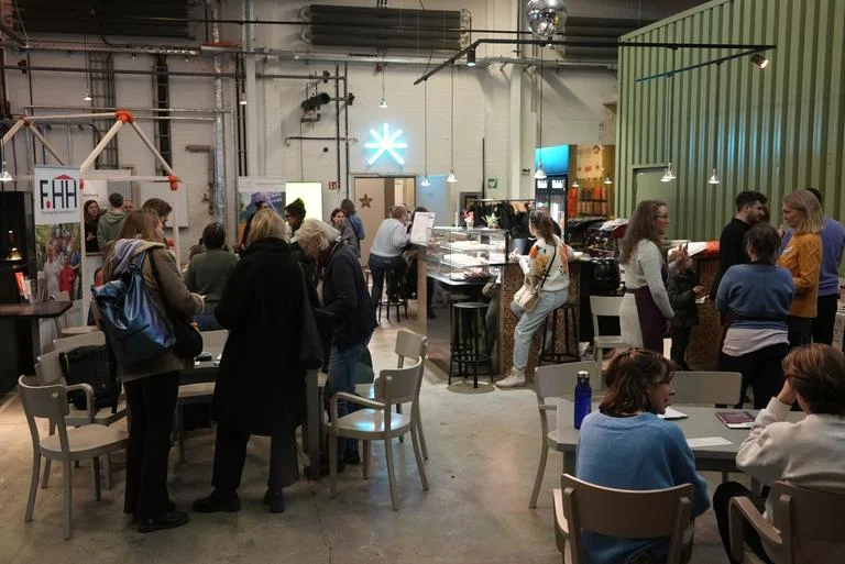 Menschen versammeln sich in einem Raum mit sitzenden und stehenden Gruppen. Im Hintergrund sind Industriewände und eine Projektionsleinwand zu sehen.