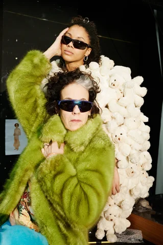 Zwei Personen posieren in avantgardistischer Kleidung. Eine trägt einen grünen Pelzmantel, die andere steht vor einer Wand aus Plüschteddys.