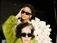 Zwei Personen posieren in avantgardistischer Kleidung. Eine trägt einen grünen Pelzmantel, die andere steht vor einer Wand aus Plüschteddys.