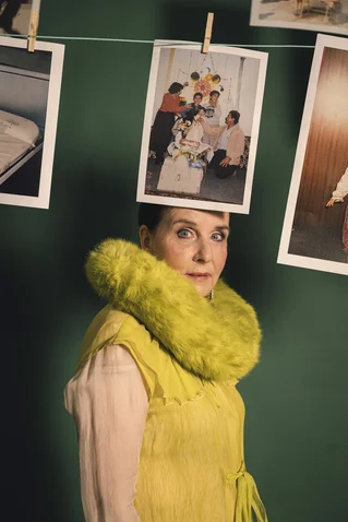 A woman in a yellow dress with a fluffy collar; photographs hang behind her on a clothesline against a green background.