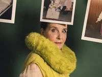 A woman in a yellow dress with a fluffy collar; photographs hang behind her on a clothesline against a green background.