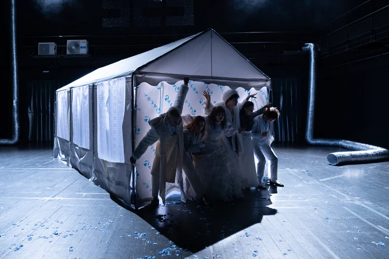 Eine Gruppe von Performern in weißen Kostümen tritt aus einem weißen Zelt auf einer dunklen Bühne hervor.