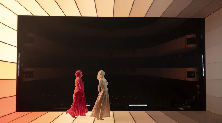 Two elegantly dressed individuals walk in front of a large, modern wall. The lighting creates a warm, inviting atmosphere.