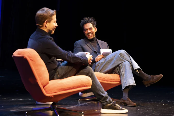 Two men sit on orange chairs on a stage, engaging in a friendly conversation. Both are dressed casually, smiling.