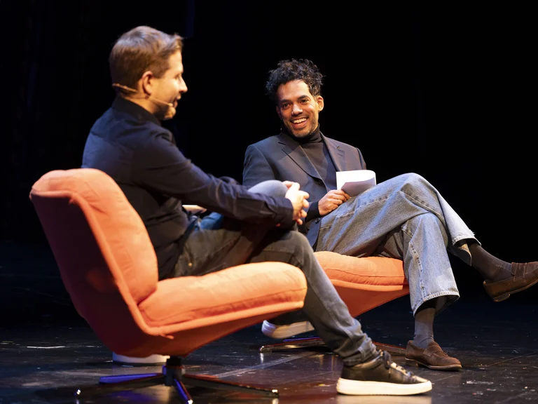 Zwei Männer sitzen auf orangefarbenen Sesseln auf einer Bühne und unterhalten sich lächelnd. Beide tragen legere Kleidung.