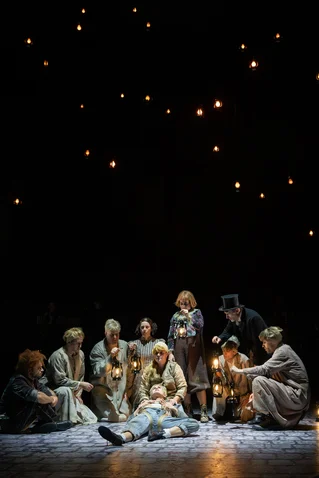 Actors in historical costumes on stage under a starry sky. One person lies on the ground, surrounded by a group with lamps.
