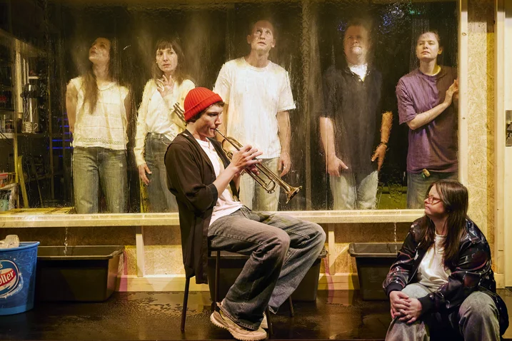 A person plays trumpet as six people watch behind a glass pane. The scene feels creative and contemplative.