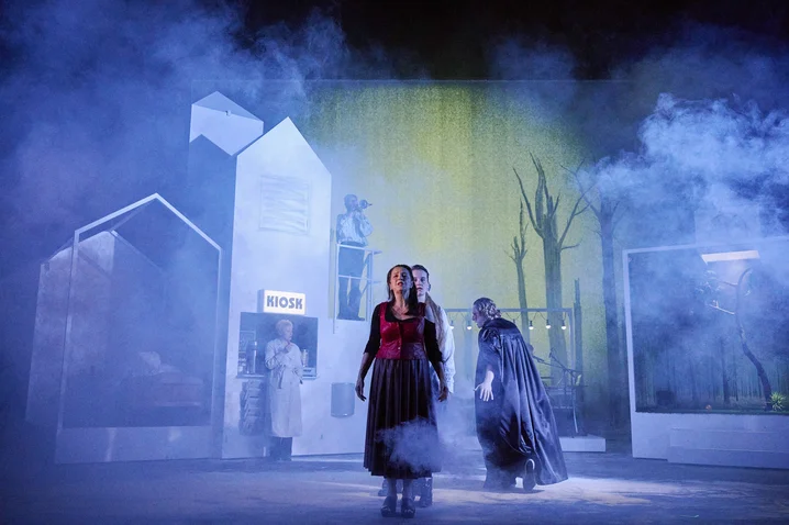 Two actors on stage in a dramatic play, surrounded by fog and minimalist sets, with a mysterious, dark backdrop.