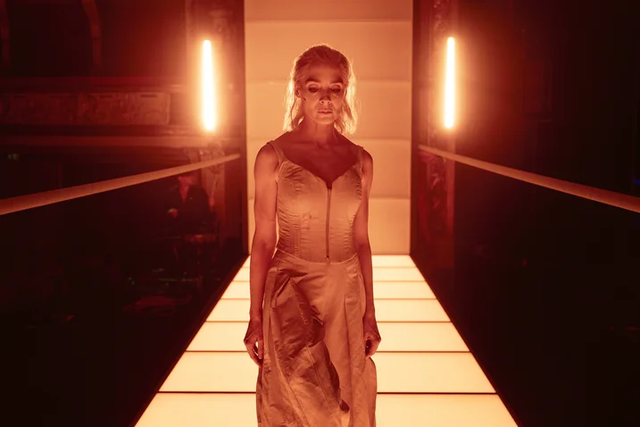 A person in an elegant dress stands on an illuminated runway with intense red lighting in the background.