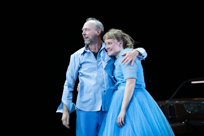 A man and a woman in blue costumes stand on stage. They embrace warmly against a dark background.