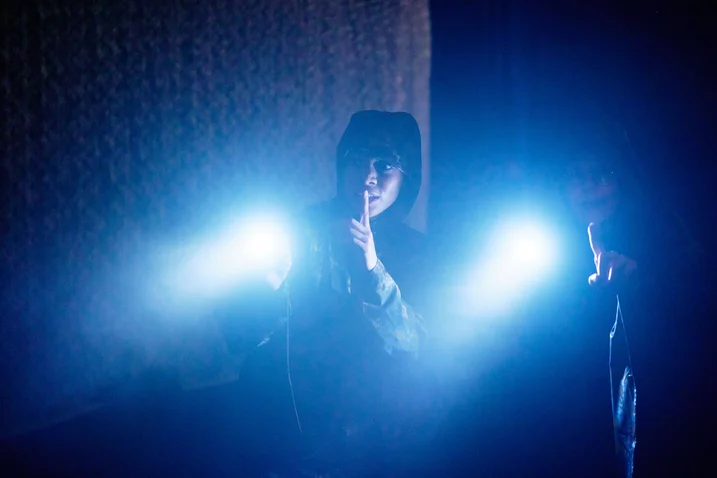 Eine Frau in einem dunklen Raum hält zwei blaue Lichter. Die Atmosphäre ist geheimnisvoll und stimmungsvoll.