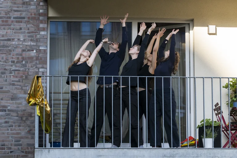 Fünf Personen in schwarzer Kleidung tanzen synchron auf einem Balkon, mit erhobenen Armen nach oben gerichtet.