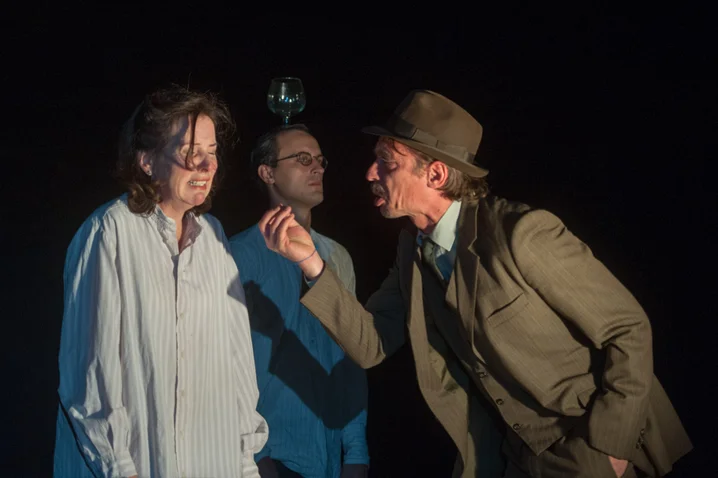 Three people perform on stage. One person in a suit speaks intently to someone in a long shirt.