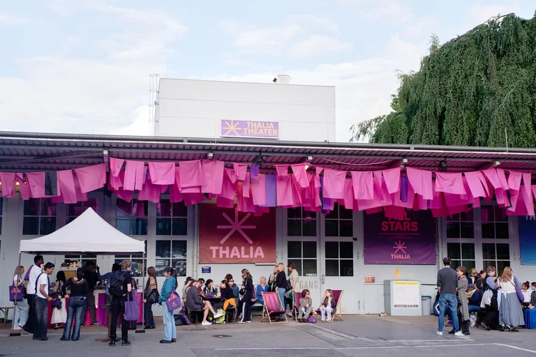 Menschen versammeln sich vor einem Gebäude mit rosa Dekorationen. Ein Zelt und Banner mit der Aufschrift "Thalia" sind zu sehen.
