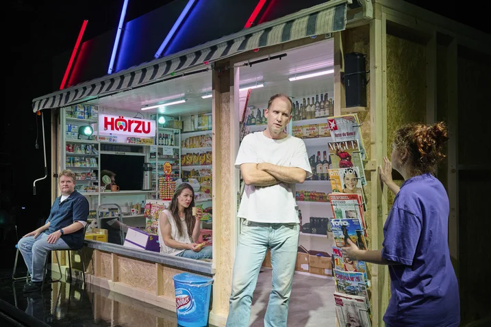 A stage with a kiosk filled with magazines and goods. Three people interact. Neon lights illuminate the scene.