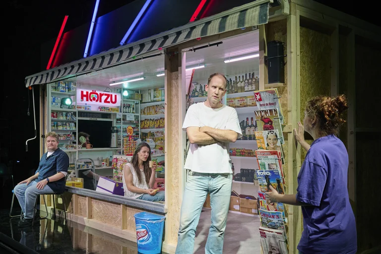 Eine Bühne mit einem Kiosk voller Magazine und Waren. Drei Personen interagieren miteinander. Neonlichter beleuchten die Szene.