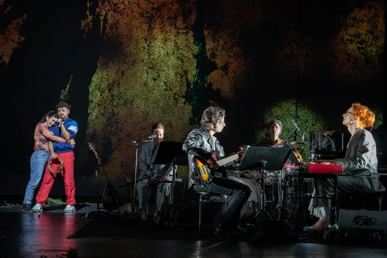 Eine musikalische Performance auf einer Bühne mit einer Band und zwei Personen, die sich innig umarmen. Hintergrund mit schummriger Beleuchtung.