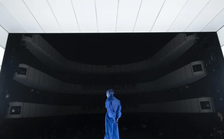 A person in a blue suit stands in a modern, dark theater against a futuristic, minimalist backdrop.