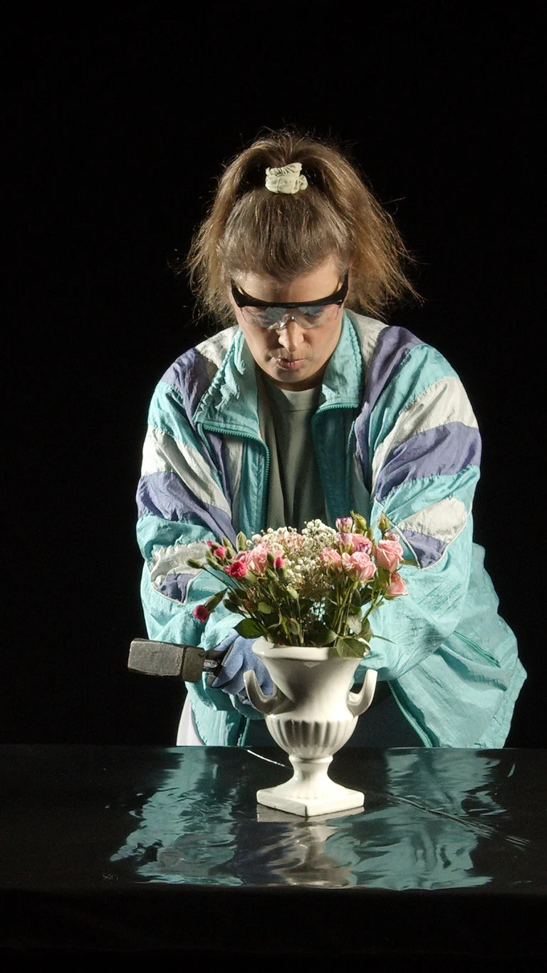 Eine Person in bunter Jacke und Schutzbrille hält einen Hammer über eine Blumenvase. Der Hintergrund ist dunkel.