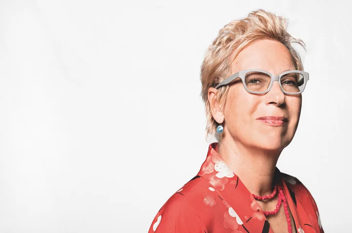 A woman with short blonde hair wears a red floral-patterned blouse and glasses, smiling gently with a calm expression.
