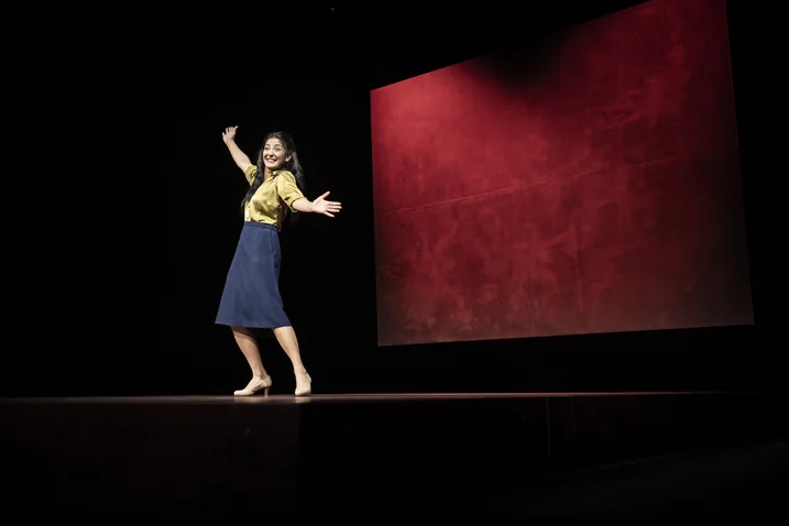A woman dances on a stage with a red background. She wears a yellow top and a blue skirt.