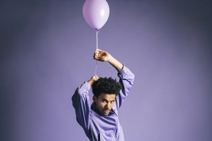 A person in a purple shirt holds a pink balloon in a dynamic pose against a purple background.