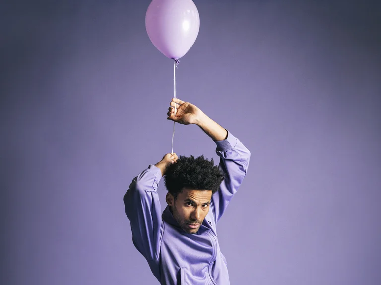 Eine Person in einem lila Hemd hält einen pinken Ballon in einer dynamischen Pose vor einem lilafarbenen Hintergrund.