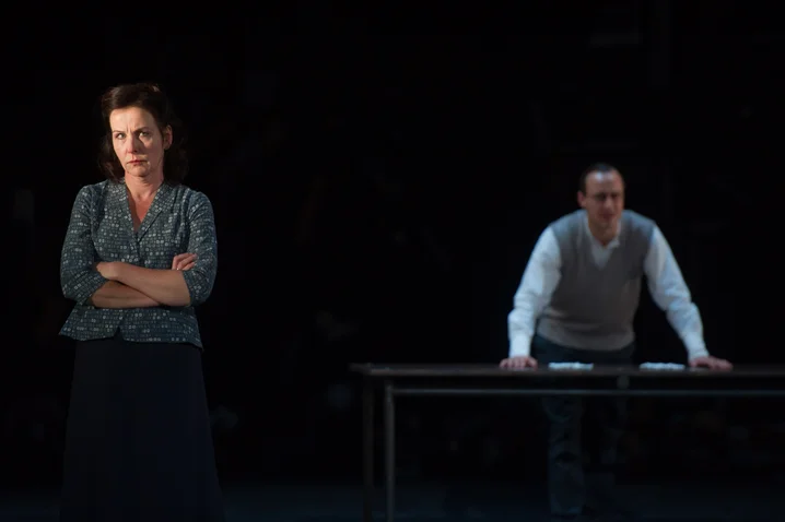 A scene on a dark stage: a woman with crossed arms stands in the foreground, a man at a table in the background.
