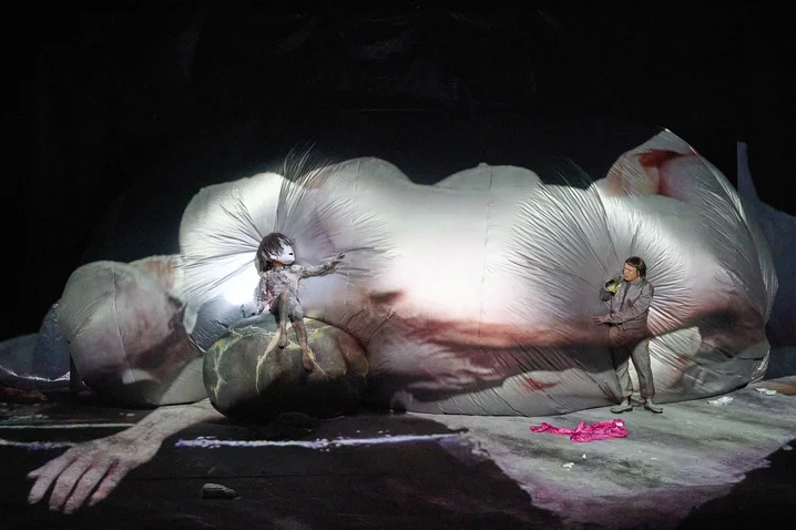Zwei Personen in futuristischen Kostümen stehen auf einer riesigen Skulptur eines liegenden Menschen, in einem theatralen Bühnenbild.
