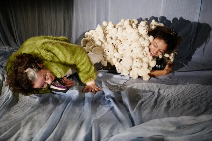 Zwei Personen liegen auf einem Bühnenboden. Eine trägt eine grüne Jacke, die andere ist in einen weißen Flaum gehüllt.