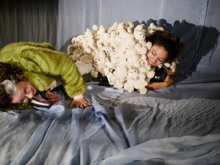 Zwei Personen liegen auf einem Bühnenboden. Eine trägt eine grüne Jacke, die andere ist in einen weißen Flaum gehüllt.