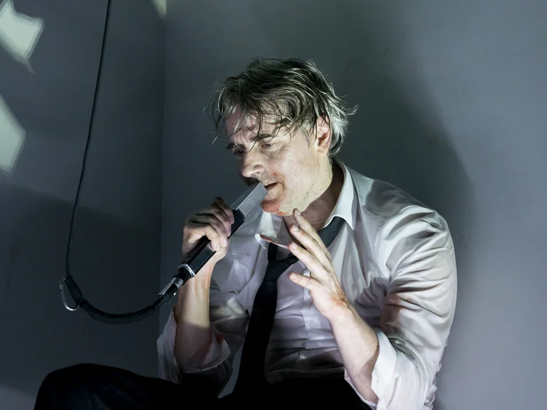 Ein Mann mit nassem Haar und zerknittertem Hemd sitzt und hält ein Mikrofon, in einem emotionalen Auftritt.