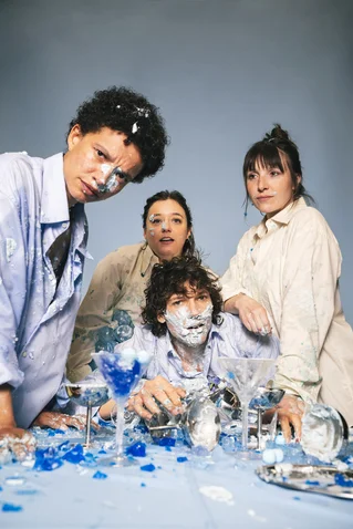 Four people wearing light clothing pose around a table with broken glass, covered in blue and white paint splatters.