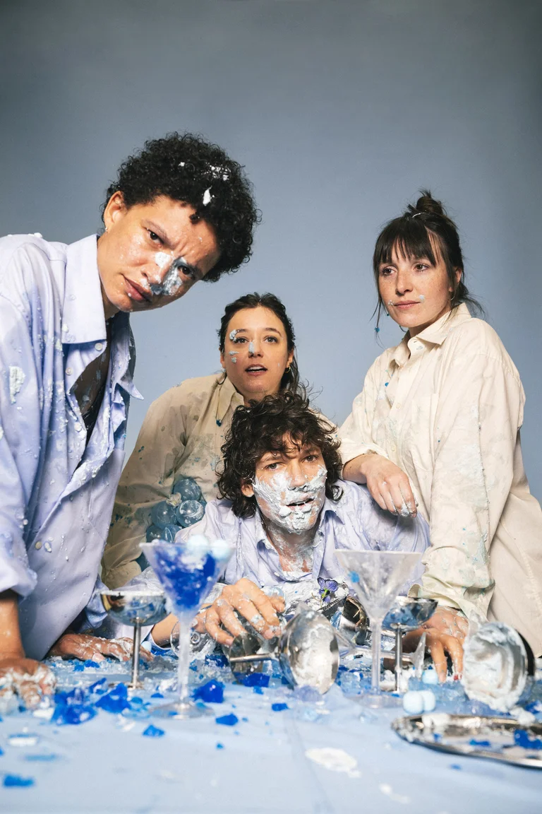 Four people wearing light clothing pose around a table with broken glass, covered in blue and white paint splatters.
