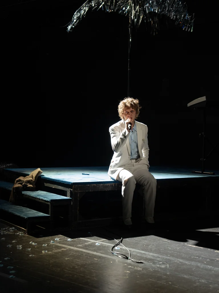 Ein Performer in einem Anzug sitzt auf einer Bühne, beleuchtet durch ein Spotlight. Eine Palme hängt leicht darüber.