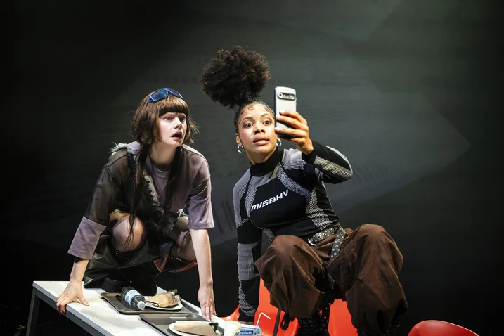 Two women are sitting on a table taking a selfie. One looks fascinated, while the other holds the phone.