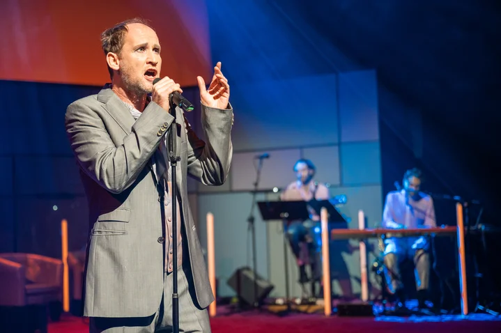A singer in a suit performs on stage. Behind him, two musicians play. The lighting is atmospheric.