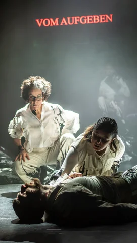 Theatrical scene with two actors leaning over a third lying on the ground. The text "VOM AUFGEBEN" glows above.