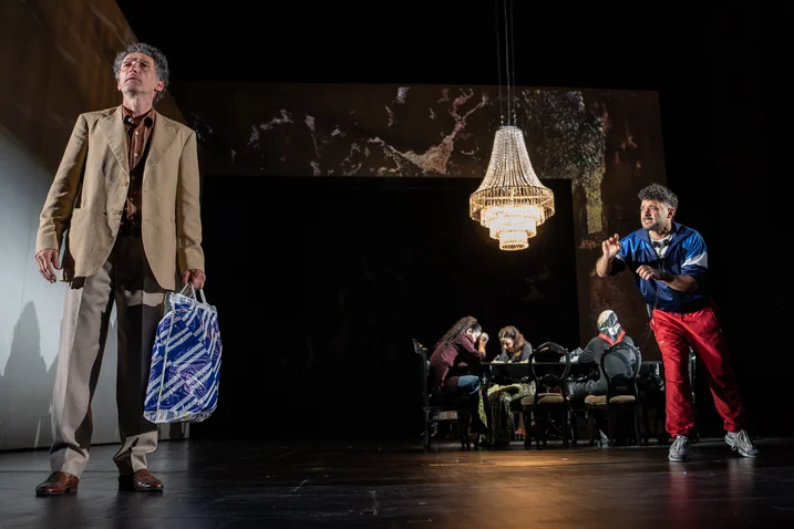 A stage scene with two performers in the foreground. One holds a bag, while a chandelier hangs in the background.